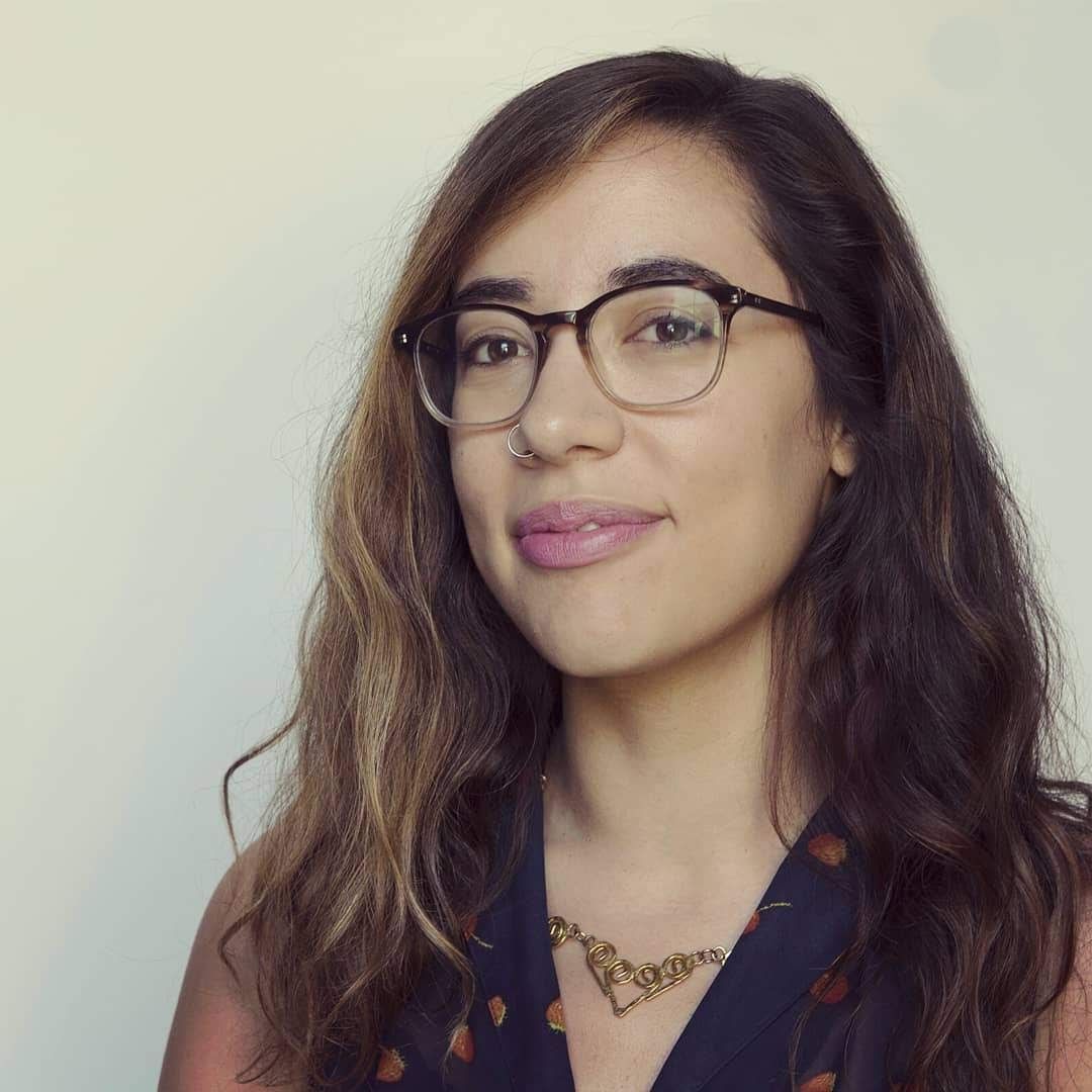 headshot of Sarah Riazati, brunette woman wearing a blue shirt and smiling against a white background